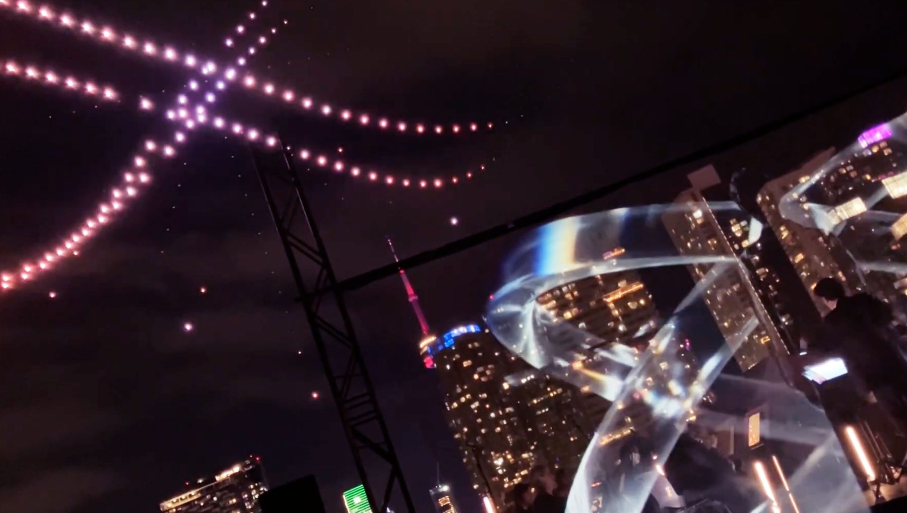 Drones fill the sky with a holographic projection against an orchestra with the skyline behind